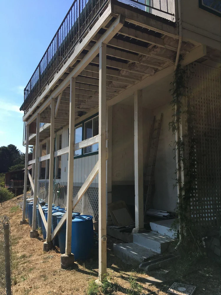 Softwood deck with more than 24 inches of clearance from the ground. Note the mesh screening draped over the gray wooden lattice beyond the deck posts to mitigate ember intrusion into the area underneath the lower portion of the structure. The blue plastic barrels were a recent purchase for a rainwater catchment; however, even if full of water, they are combustible and should not be stored near structures.